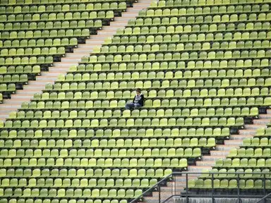 An empty stadium's baseball box seats, where a man sits on a bench, gazing out at the field