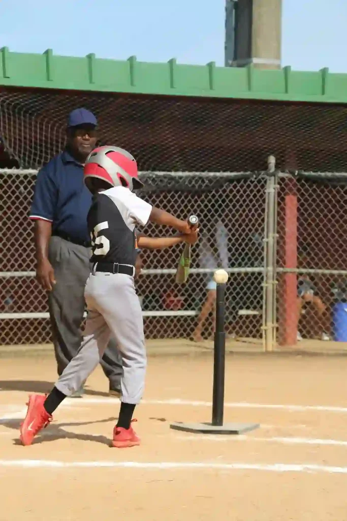 What Is a Utility Player in Baseball- Everything You Need To Know 2 A young baseball player in red gear, illustrating what is a utility player in baseball, prepares to hit a ball on a tee at a sunny field, with an umpire observing nearby.