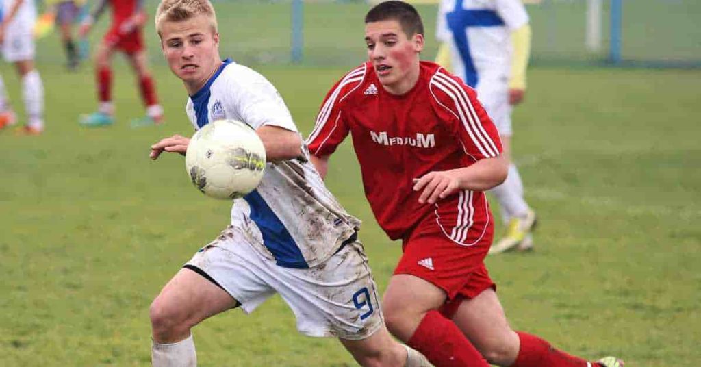 What Does 1st and 15 Mean in Football? A Football Guide 2 Two young male soccer players compete for the ball on a grassy field, one in a muddy white jersey and the other in a red jersey, illustrating the intensity of the game much like understanding what 1st and 15 mean in football.