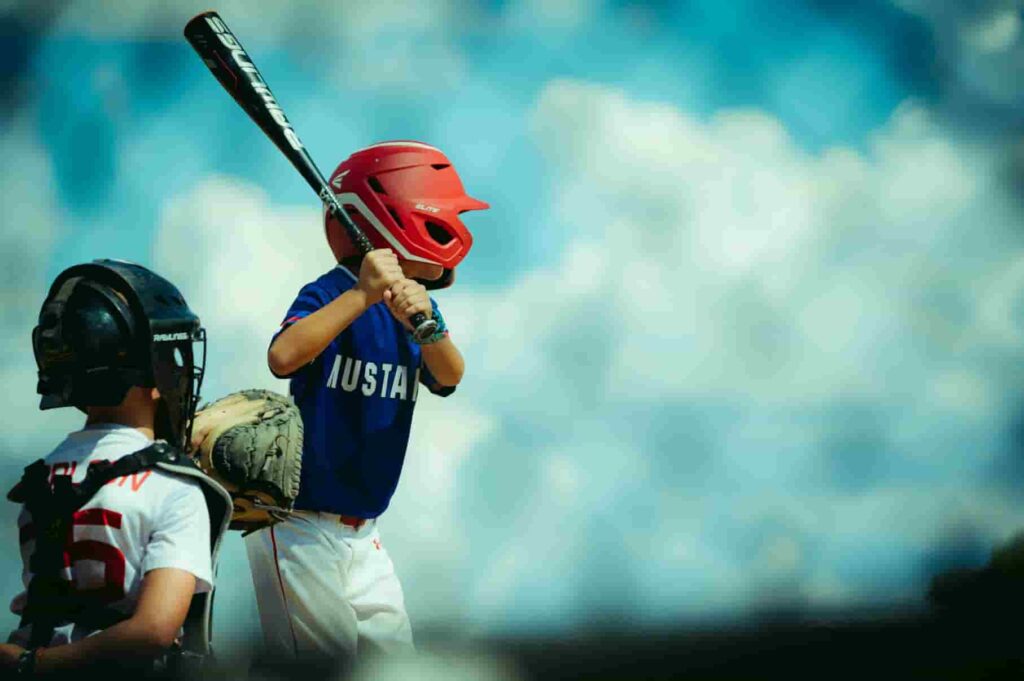 During one of the high school baseball games, a young baseball player in a red helmet stands at the plate, bat poised, while a catcher in black gear prepares to receive the pitch.
