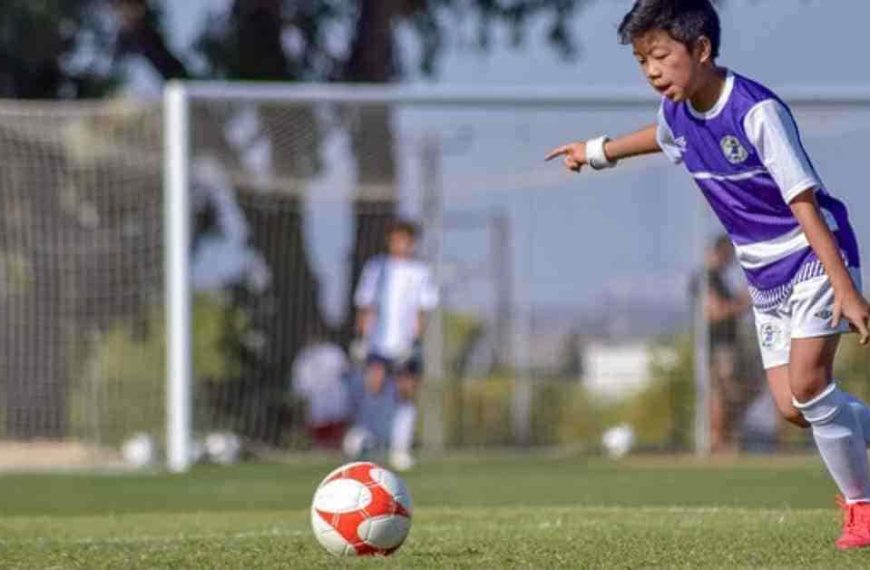 A young boy wearing a football helmet with stickers kicking a ball on a field.