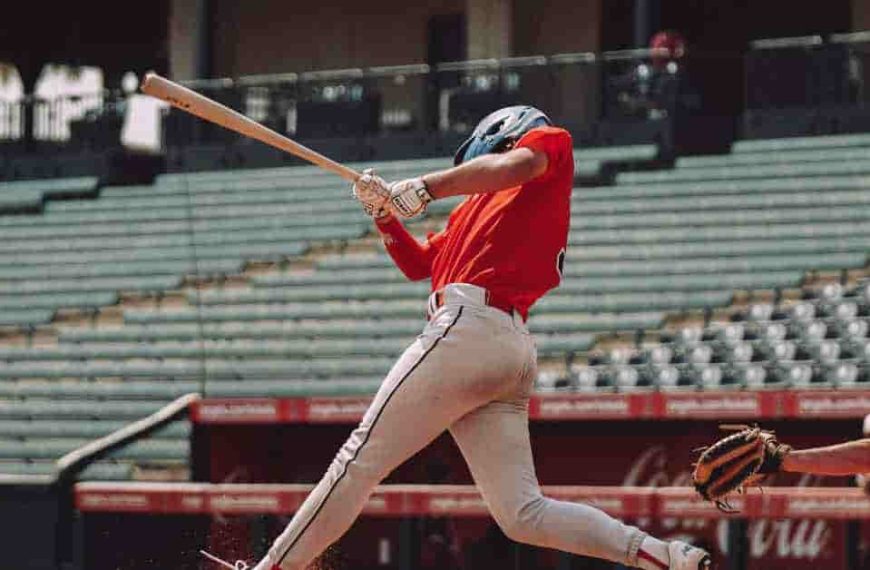 Baseball player wearing baseball cleats and swinging a bat on top of a field.