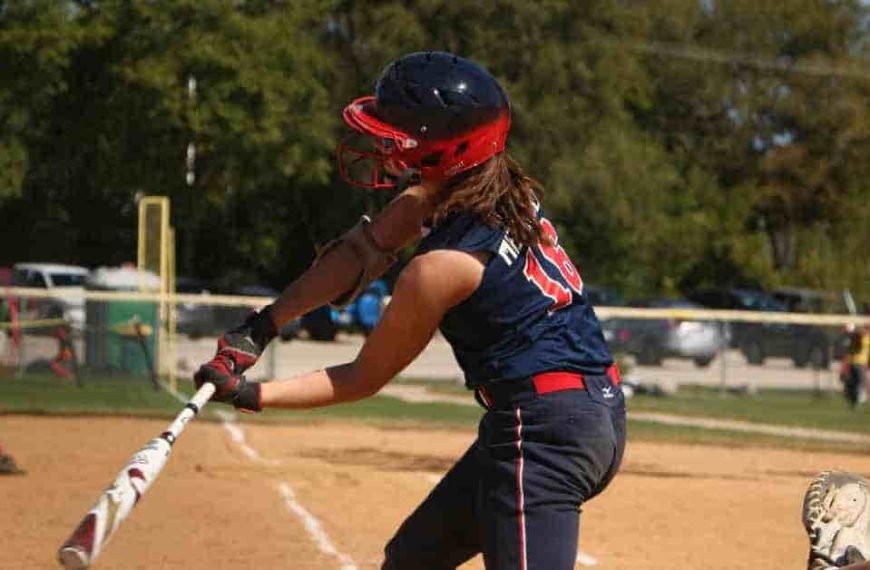 A girl swinging a baseball bat on a baseball field, with a radar gun nearby to measure pitch speed