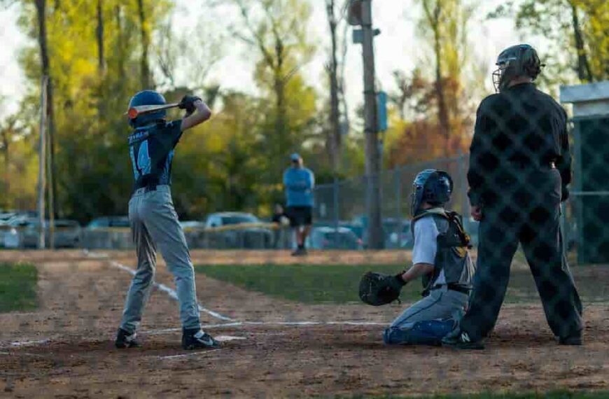 A young player in the baseball college uniforms stands at bat while the catcher and umpire are ready. In the background, a coach observes the game from near the fence.