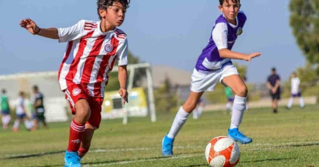 Two youth soccer players in action on the field, one in red and white, the other in purple, with a backdrop that could lead to discussions on topics like how long is a football game.