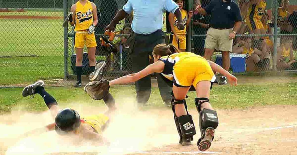 Bat Rolling.. What Is It, Benefits, Risks, and How It Works 3 A player slides into home plate during a tense moment in a softball game while an opposing catcher prepares to tag her out, highlighting the competitive edge gained through bat rolling.