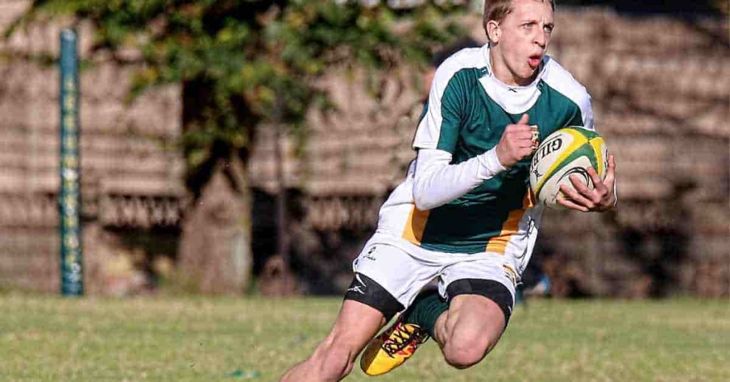 How should football cleat fit?: Finding The Perfect Fit 2 A rugby player in green and white gear sprints across a grassy field, holding a rugby ball, wearing well-fitted cleats that demonstrate how should football cleat fit for optimal performance and support.