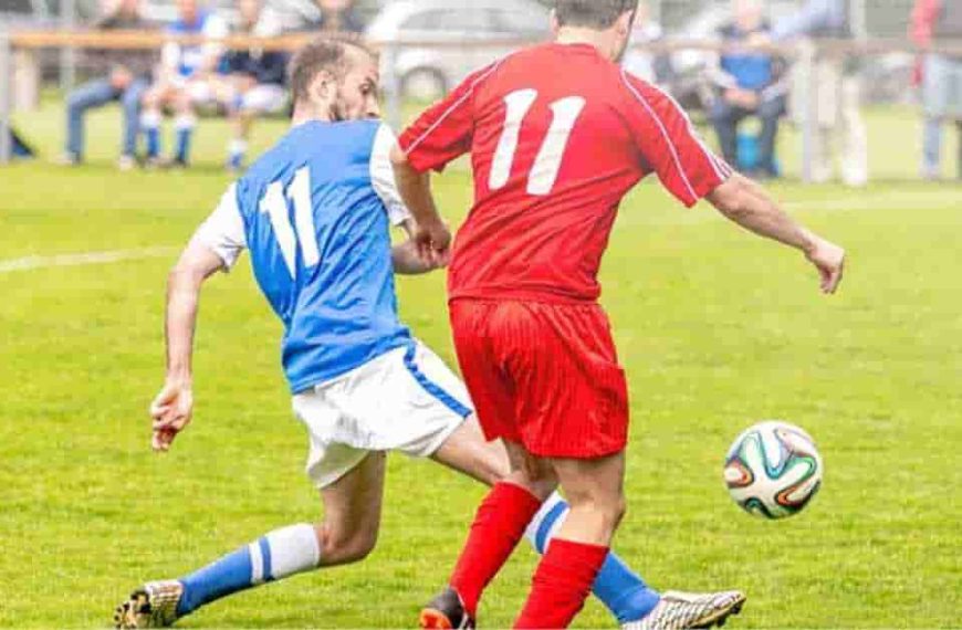 A couple of men playing a game of soccer on a grassy field, with one player kicking the ball and the other preparing to defend. The game details are tbd in football.